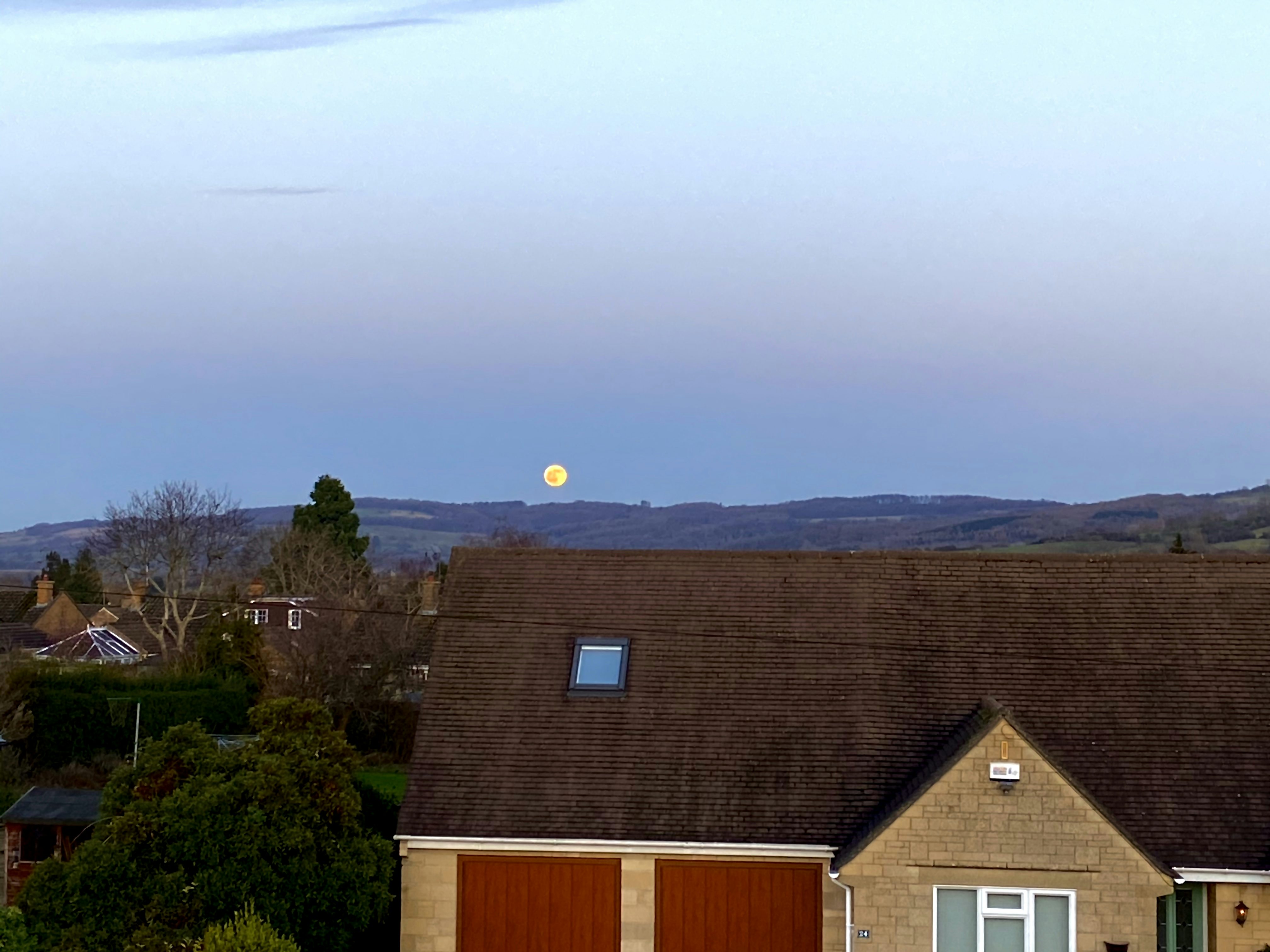 A very bright, orange moon rises up over some houses and the hills of Gloucestershire