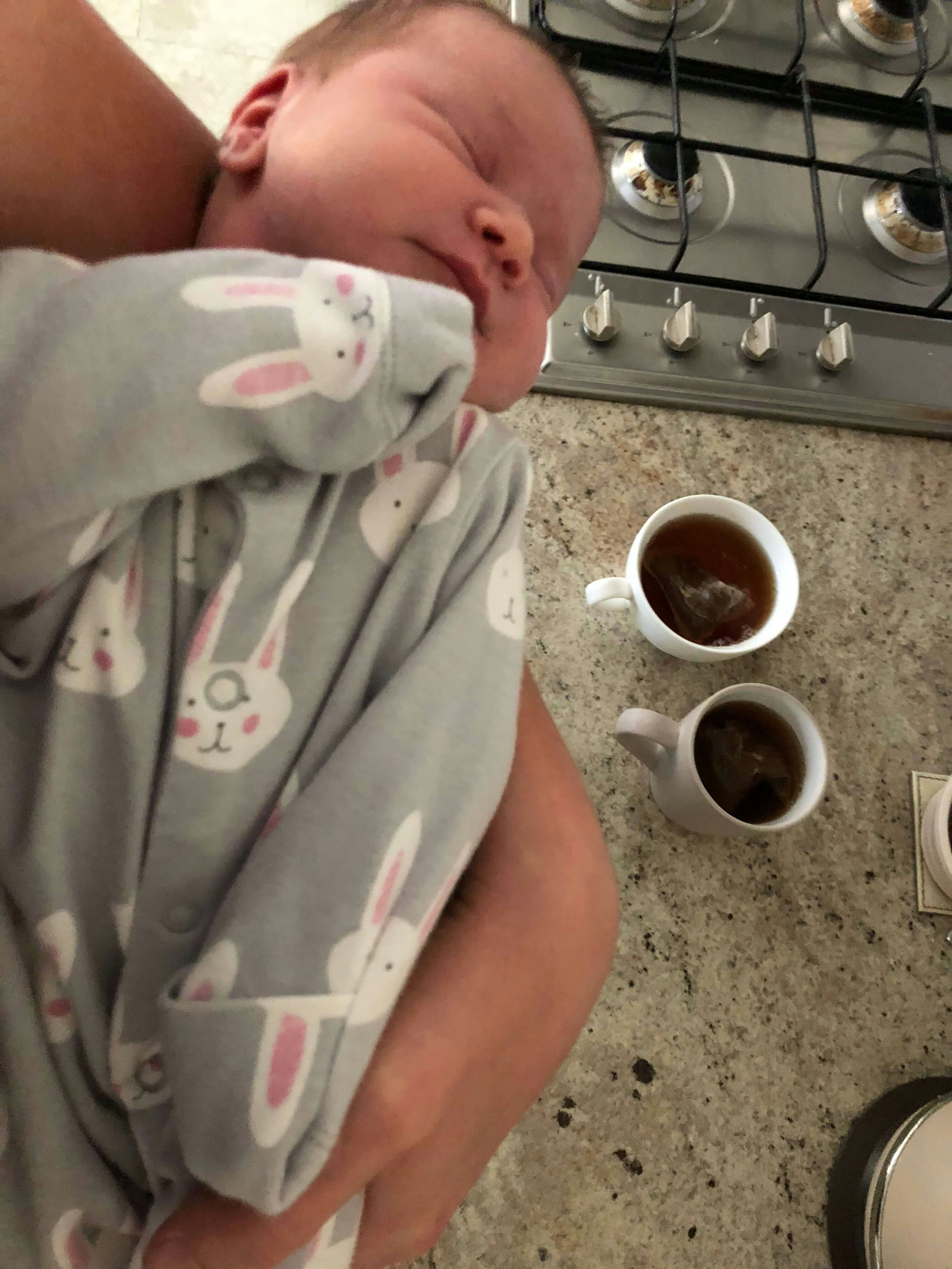 A baby is cradled while two cups of tea are brewed in the background