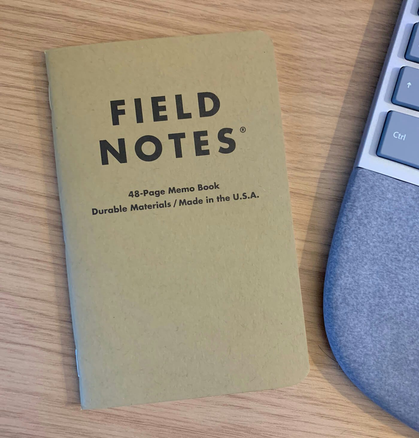 A fresh Field Notes notepad sits on a brown desk, next to a keyboard